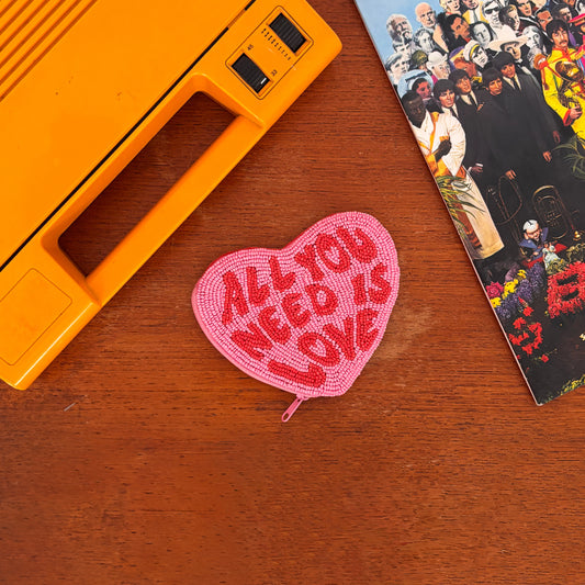 A music-themed accessory, this pink Beatles heart-shaped purse with ALL YOU NEED IS LOVE in red rests on a wooden surface beside an orange device and a colourful album cover.