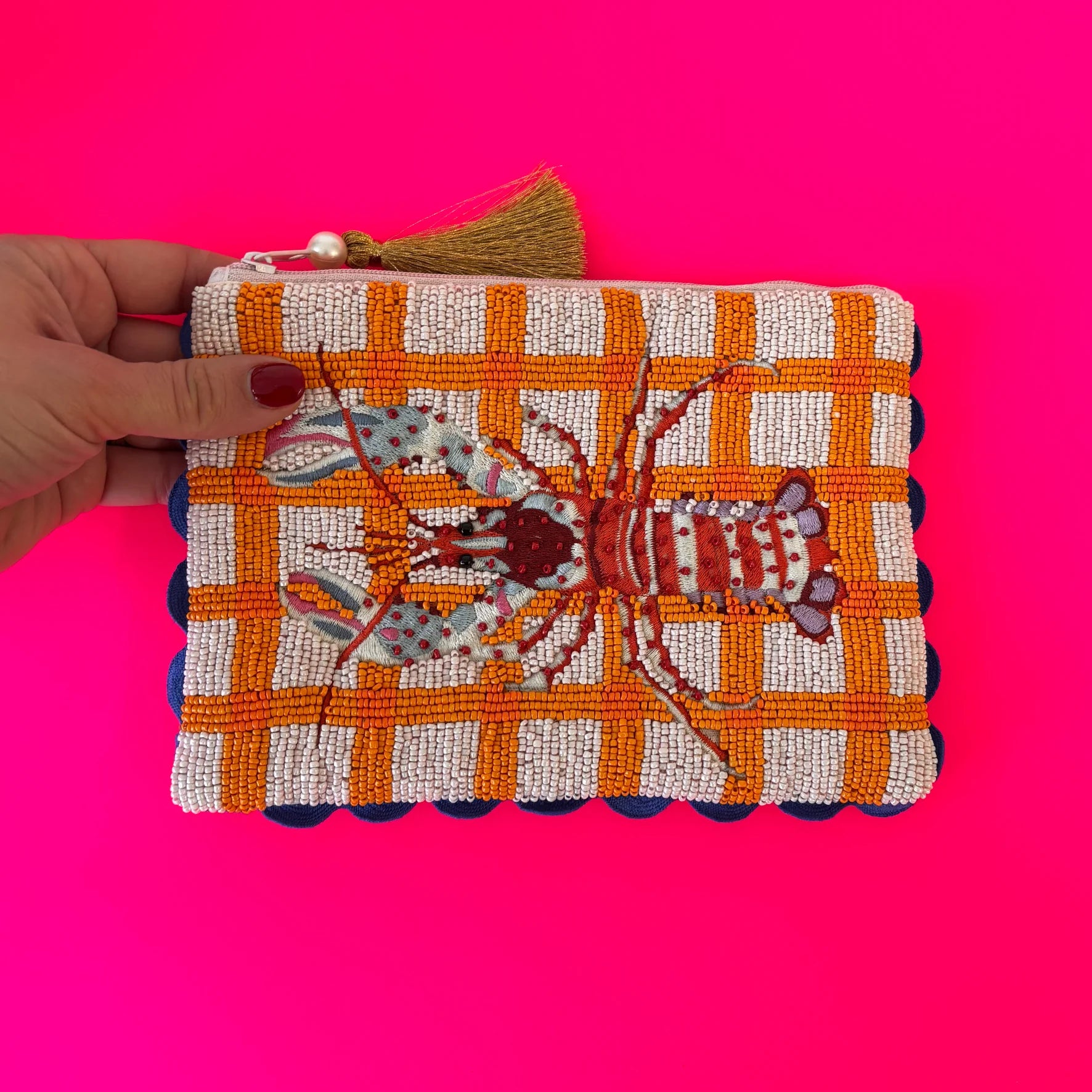 A hand holds a vegan-friendly beaded pouch featuring an orange and white chequered pattern with a beaded lobster design on the front, set against a bright pink background.