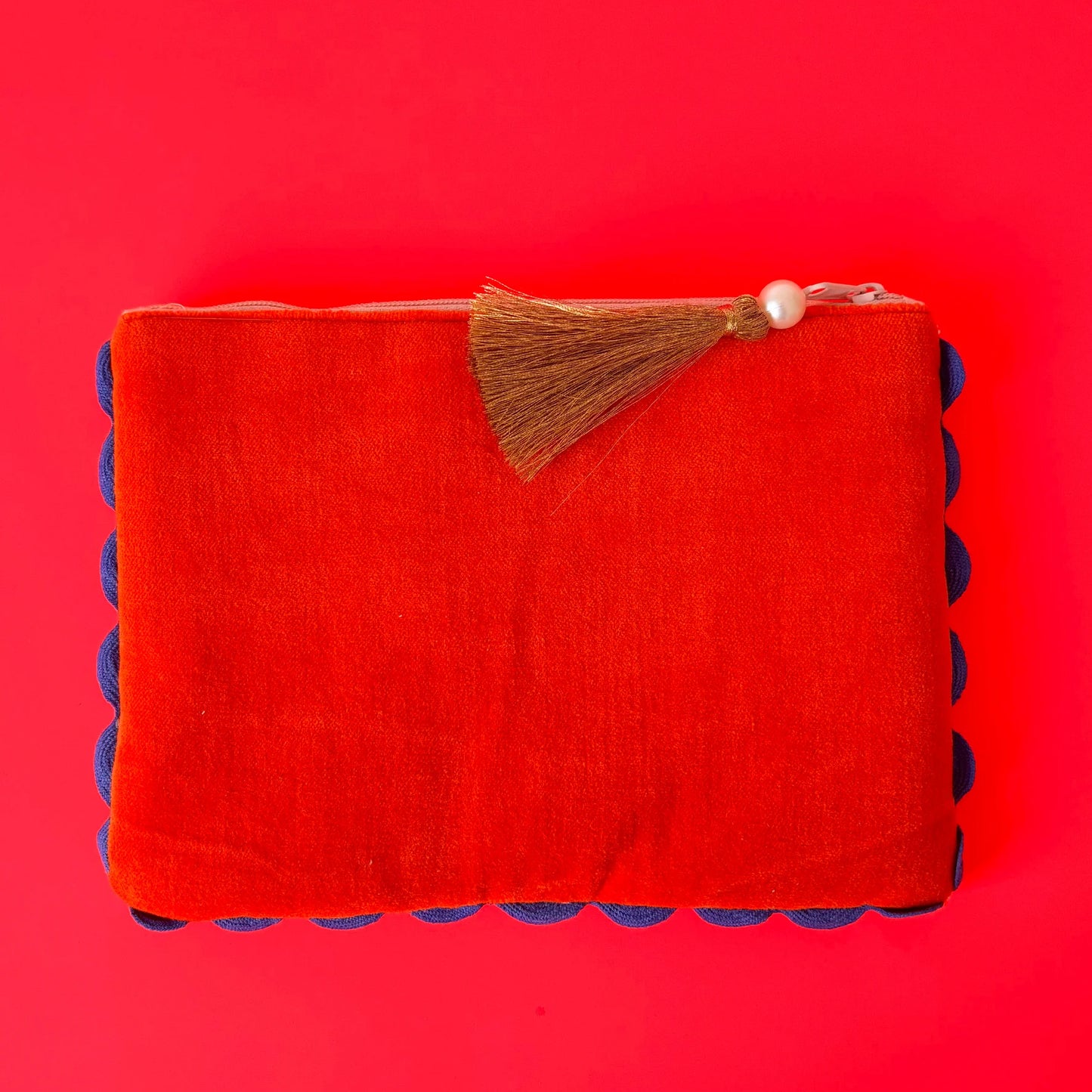 A rectangular, hand-crafted red fabric pouch with a zip and brown tassel, adorned with blue pom-pom trim along the sides, set against a solid, bright red background.