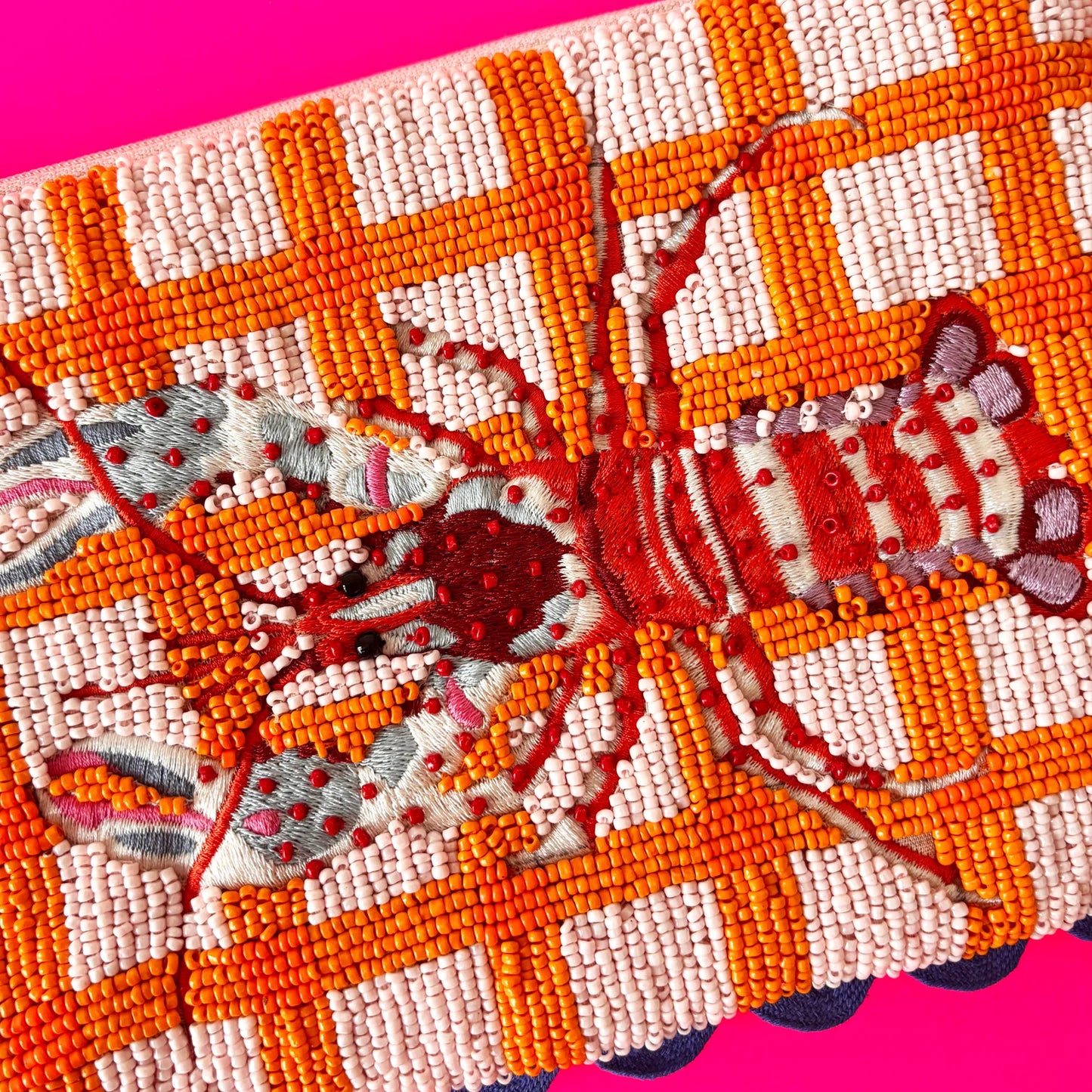 Beaded embroidery of a red lobster on an orange and pink chequered background, placed on a bright pink surface—a vegan-friendly accessory and uniquely hand-crafted pouch.