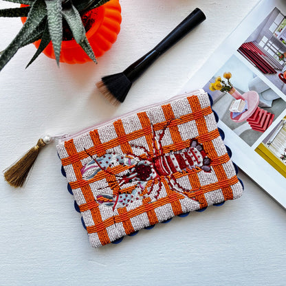 Beaded pouch featuring a lobster motif on an orange and white chequered background; this vegan-friendly accessory rests beside a make-up brush, a plant, and an open magazine on a white surface.