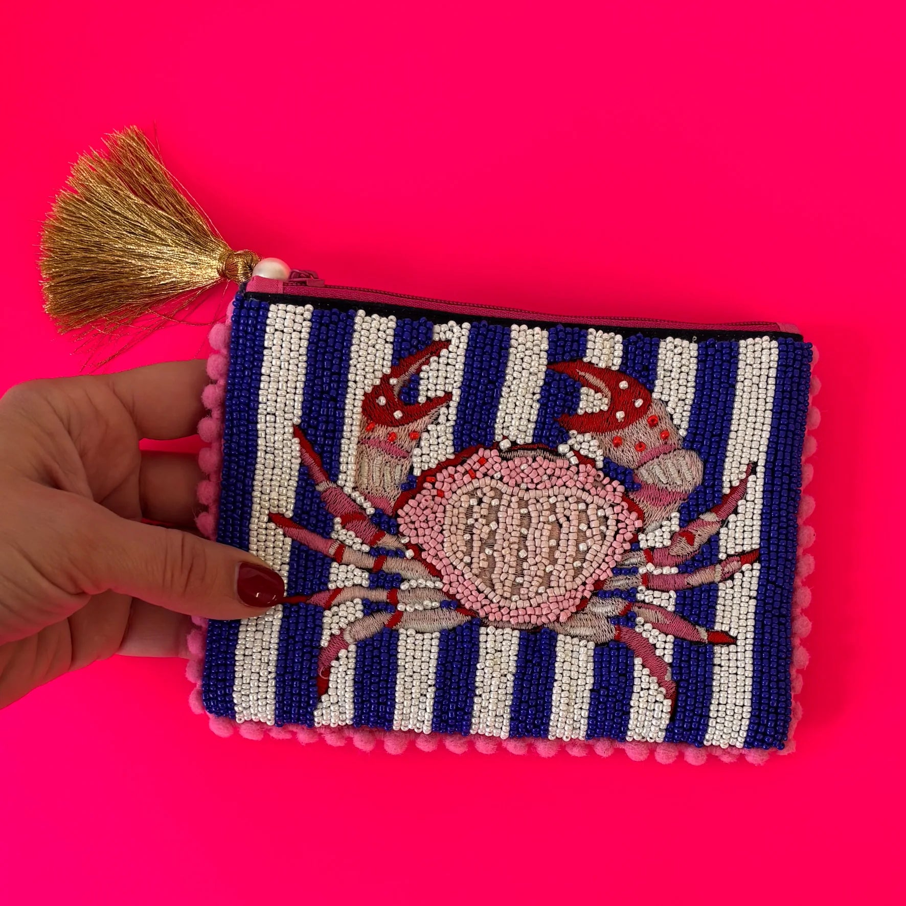 A hand holds a handcrafted beaded pouch featuring blue and white nautical stripes, a red crab motif, pink pom-pom trim, and a gold tassel, set against a vivid pink background.