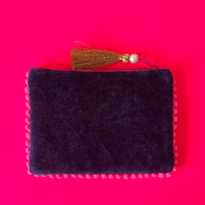 A rectangular, dark purple handcrafted pouch with pink edging and a gold tassel zip pull, set on a vivid pink background.