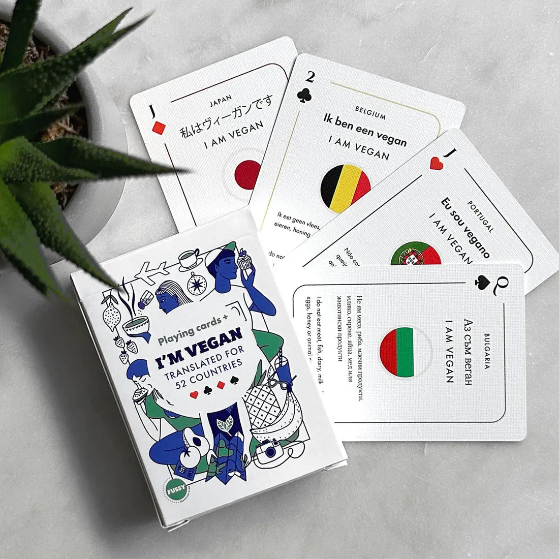 A deck of Vegan Travel Playing Cards rests on a table, each card featuring "I am vegan" translated into various languages alongside their respective national flags—an ideal accessory for vegan travellers.