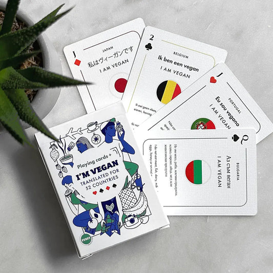 A deck of Vegan Travel Playing Cards rests on a table, each card featuring "I am vegan" translated into various languages alongside their respective national flags—an ideal accessory for vegan travellers.