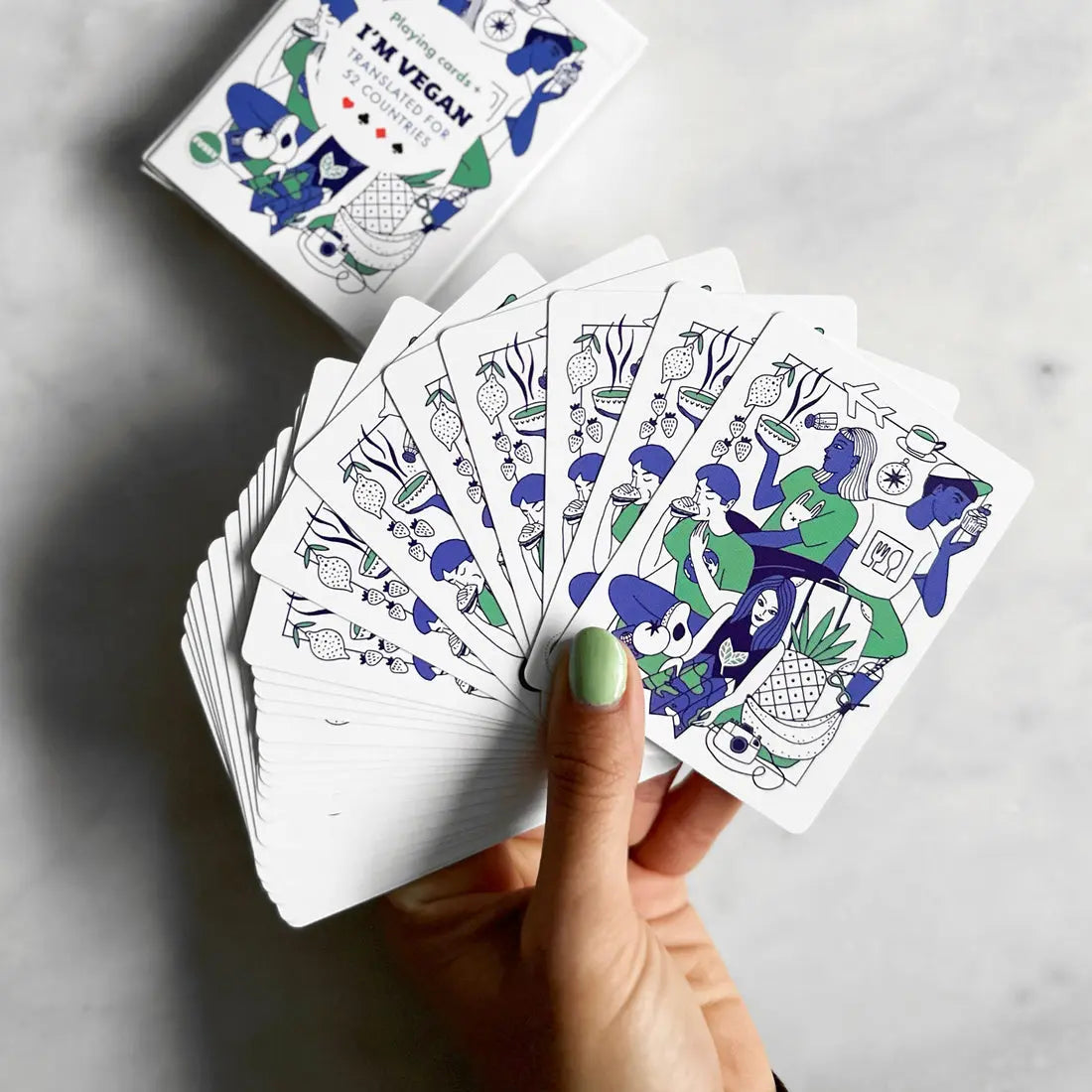 A hand holds a fanned-out deck of Vegan Travel Playing Cards featuring vegan-themed designs; ideal for vegan travellers, with the card box marked IM VEGAN! visible in the background.