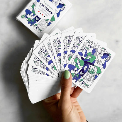 A hand holds a fanned-out deck of Vegan Travel Playing Cards featuring vegan-themed designs; ideal for vegan travellers, with the card box marked IM VEGAN! visible in the background.