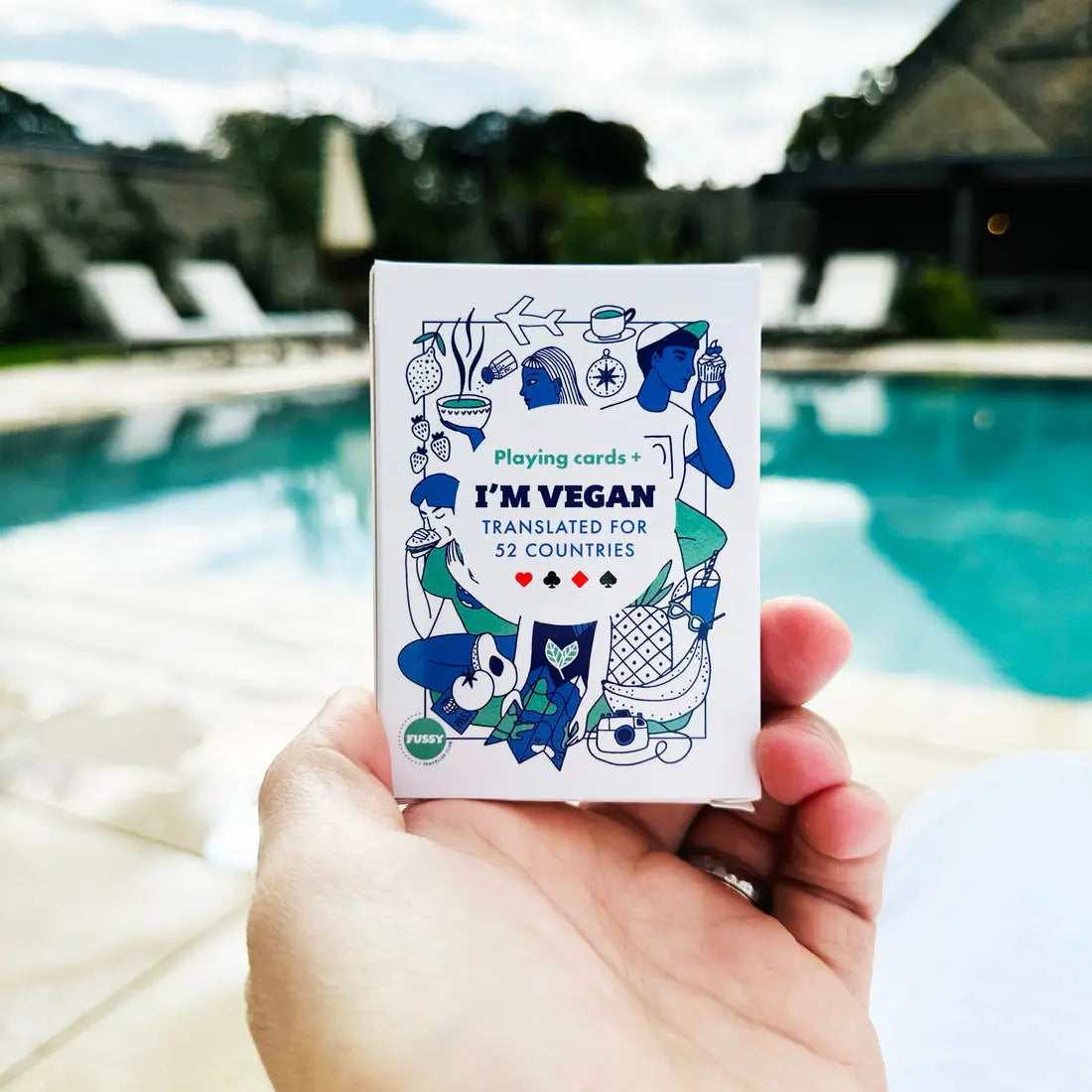 A hand holds Vegan Travel Playing Cards labelled IM VEGAN: Translated for 52 Countries, ideal for vegan travellers, with a swimming pool and lounge chairs visible in the background.