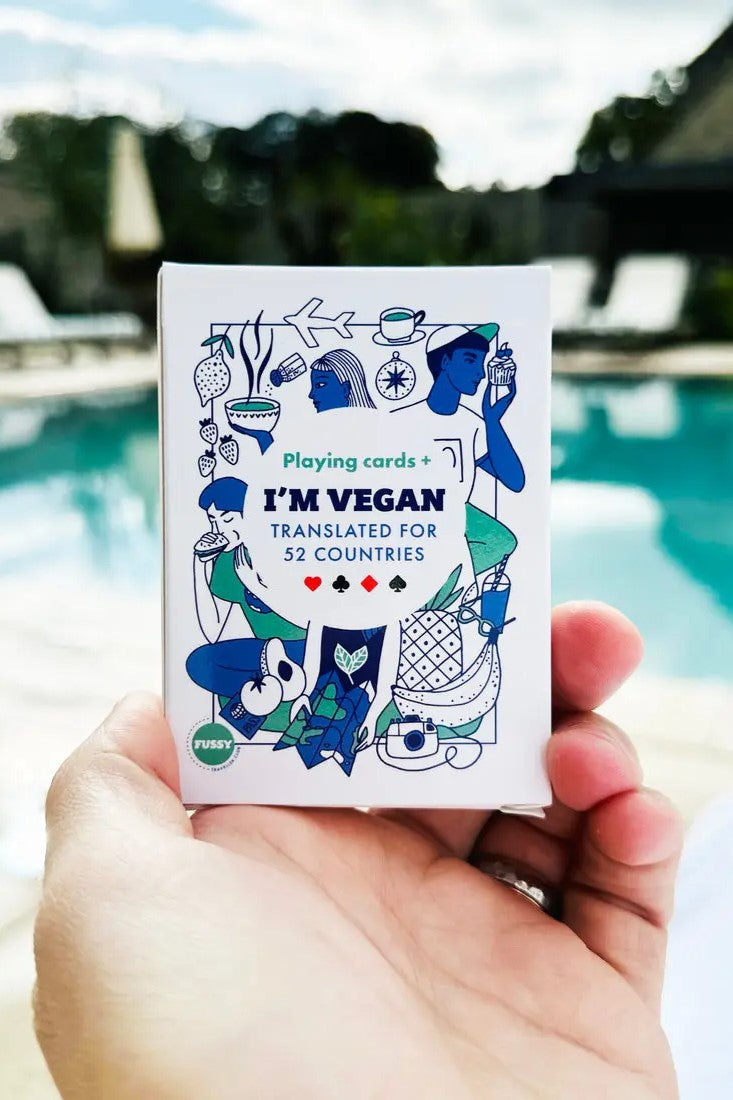 A hand holds Vegan Travel Playing Cards beside the pool—language translation cards for vegan travellers, with colourful illustrations and translations for 52 countries.