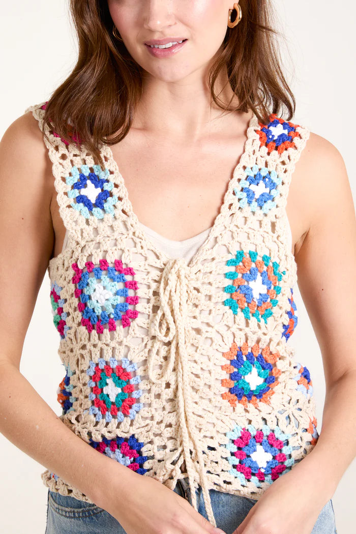 A fashion-forward woman wears a vibrant, colourful patchwork crochet vest top over a crisp white shirt, perfectly complemented by her classic blue jeans.