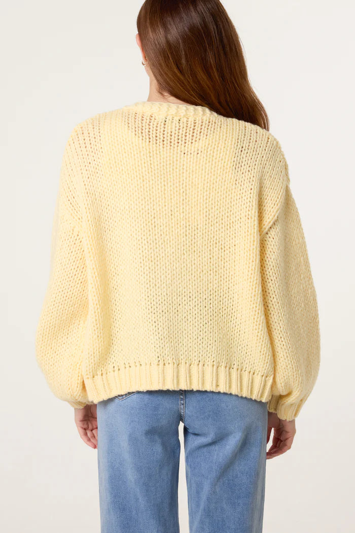 Woman with long brown hair wearing a loose, chunky yellow knitted jumper with subtle floral embroidery and blue jeans, seen from the back against a plain background.