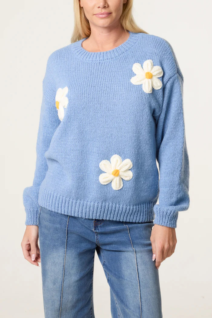 A person wearing a light blue Flocking Daisy Knitted Jumper featuring large white and yellow daisy appliqués, crafted from sustainable materials, stylishly paired with blue jeans.