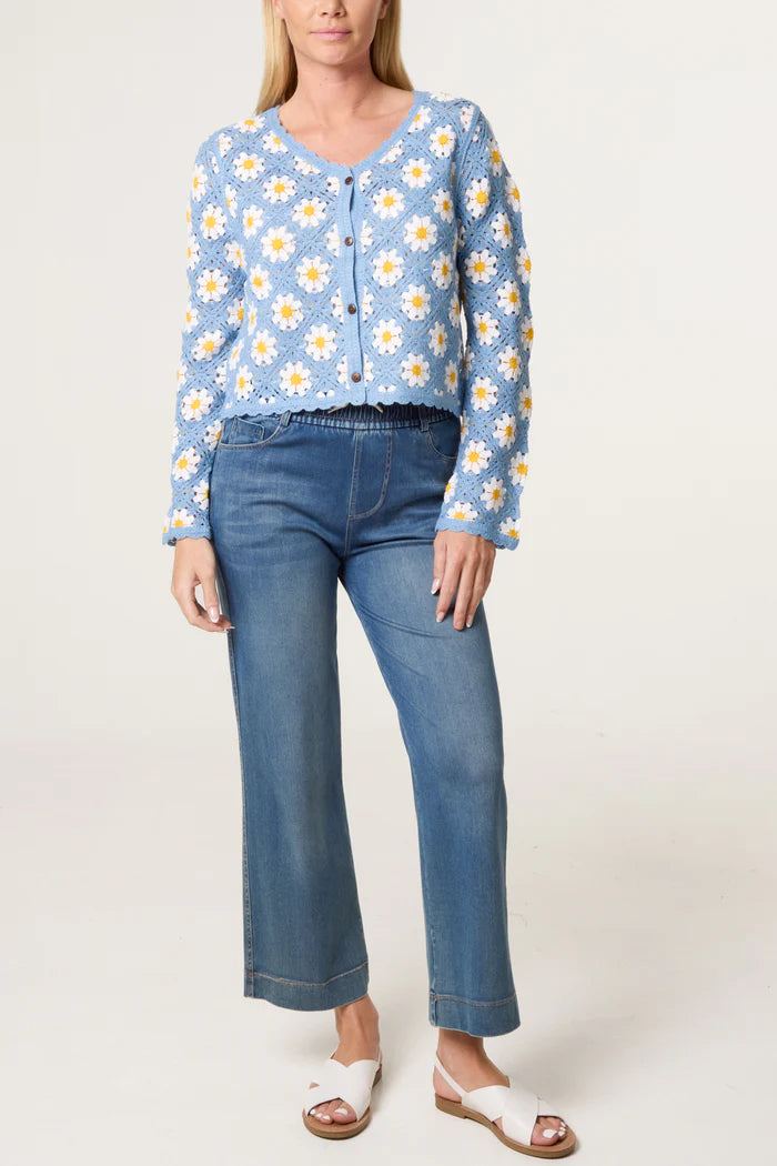 A person wearing a vintage-style light blue crochet cardigan featuring white and yellow daisy motifs, blue jeans, and white sandals stands against a plain background.
