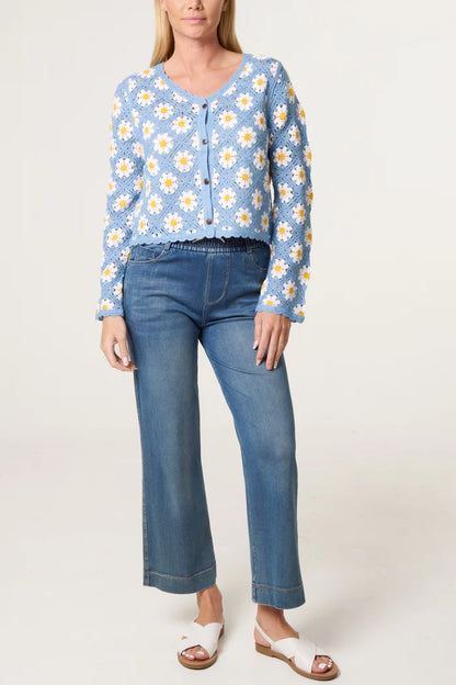 A person wearing a vintage-style light blue crochet cardigan featuring white and yellow daisy motifs, blue jeans, and white sandals stands against a plain background.