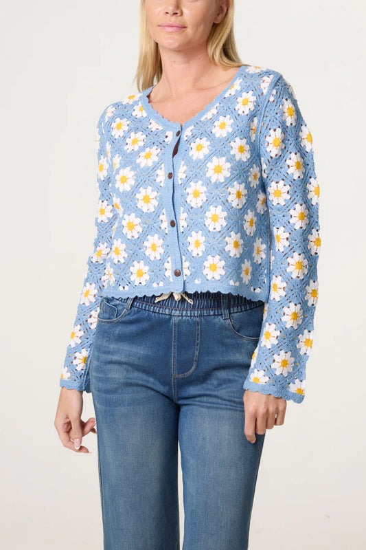A person wearing a vintage-inspired blue crochet cardigan adorned with white and yellow daisy motifs, paired with blue jeans, stands against a plain backdrop.