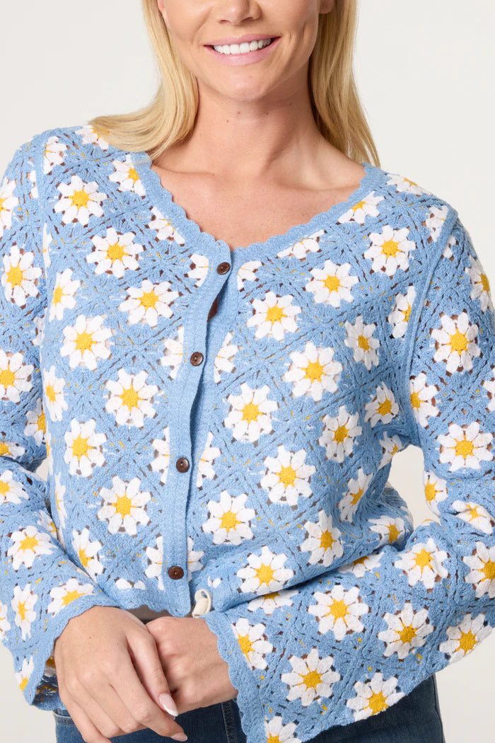 Woman wearing a blue crocheted cardigan with white and yellow daisy motif, brown buttons, and scalloped edges, smiling against a light background.