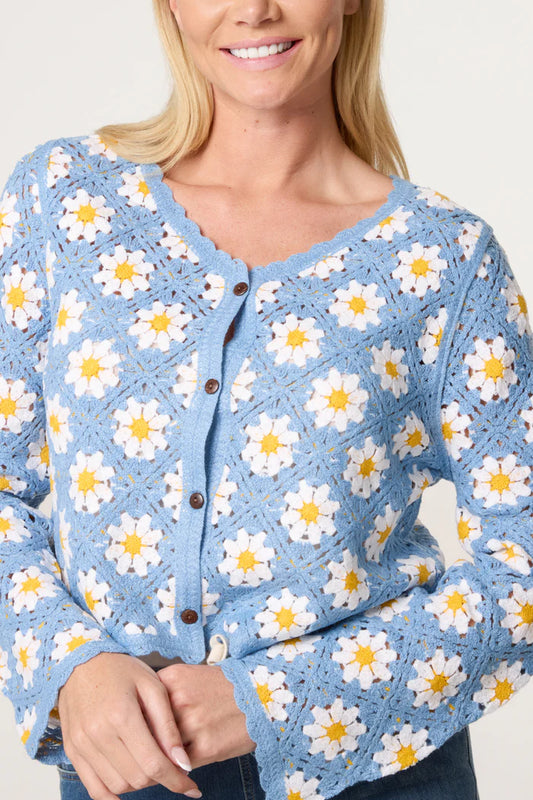 Woman wearing a blue crocheted cardigan with white and yellow daisy motif, brown buttons, and scalloped edges, smiling against a light background.
