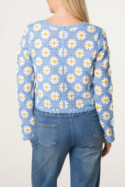 A person wearing a vintage-inspired crochet cardigan featuring white and yellow daisy motifs, paired with blue jeans, seen from the back.