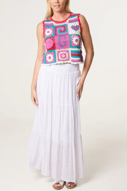 A woman in a colourful, 70s-inspired crocheted sleeveless top and a long, white tiered skirt stands against a simple light background.