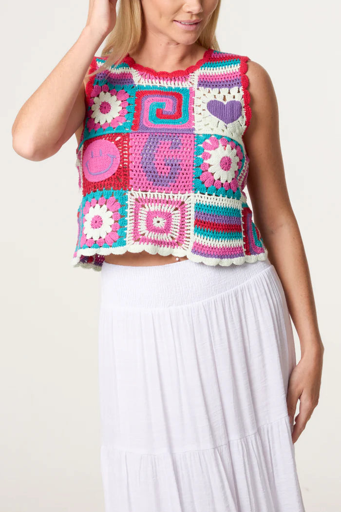 A person sporting a colourful retro patchwork crochet vest adorned with assorted patterns and symbols, teamed with a white skirt, stands before a plain background, radiating vintage charm.