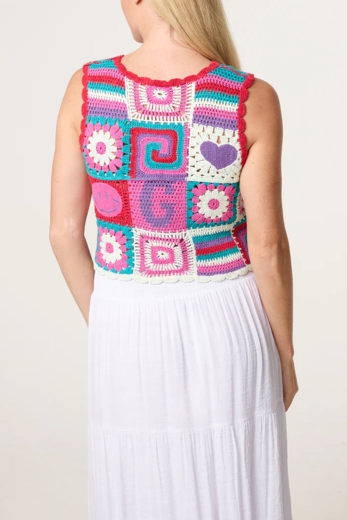 A woman with long blonde hair is seen from behind wearing a retro patchwork crochet vest adorned with geometric and floral patterns over a white skirt, evoking vintage charm.