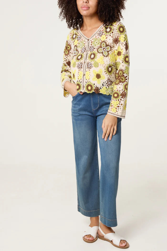 A person wearing a floral V-neck blouse, blue wide-leg jeans, a lightweight crochet jumper, and white sandals stands against a plain background.