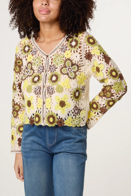 Person wearing a lightweight crocheted jumper with yellow, brown, and green floral patterns and blue jeans, standing against a plain background.