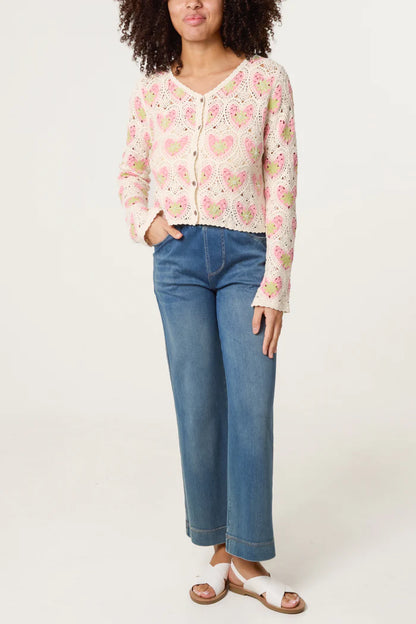 A person in a pink and white heart-patterned handmade crochet cardigan, blue wide-leg jeans, and white sandals stands against a plain backdrop.
