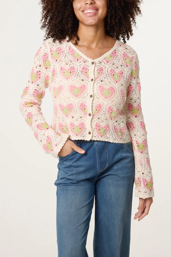 A person wears a handmade crochet cardigan featuring pink and green heart patterns, paired with high-waisted blue jeans, standing against a plain background.