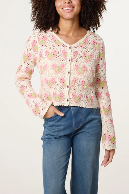 A person wears a handmade crochet cardigan featuring pink and green heart patterns, paired with high-waisted blue jeans, standing against a plain background.