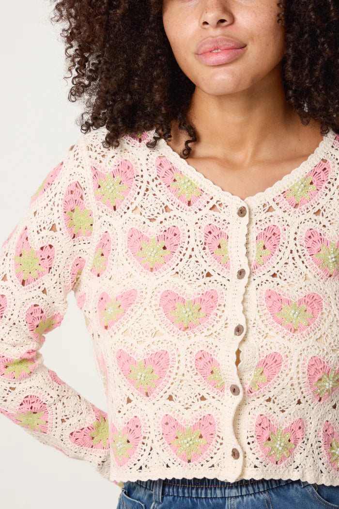 A woman wears a cream-coloured handmade cardigan featuring pink and green heart patterns, fastened with buttons down the front.