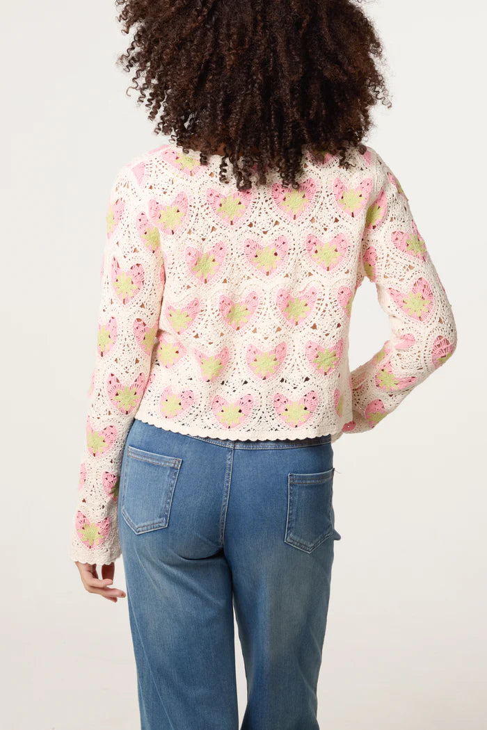 A person with curly hair is seen from behind, wearing a pink and white handmade crochet cardigan featuring heart patterns and blue jeans.