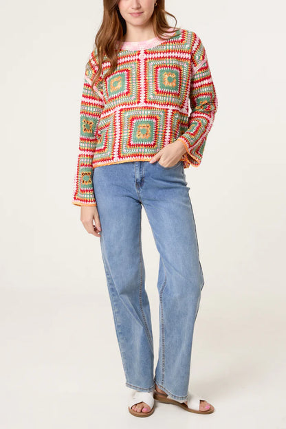 A person wearing a vibrant patchwork crochet jumper, light blue jeans, and white sandals stands against a plain backdrop.