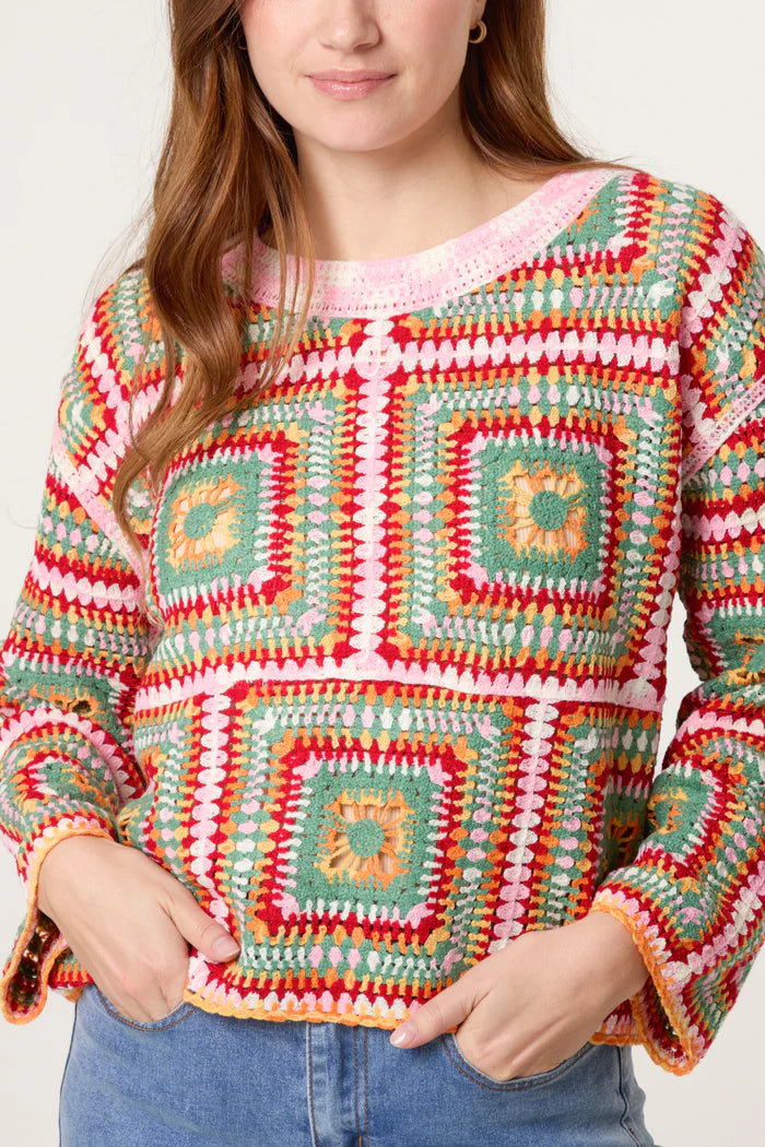 A person wearing a vibrant patchwork crochet jumper featuring geometric square patterns in red, green, yellow, and pink, paired with blue jeans.