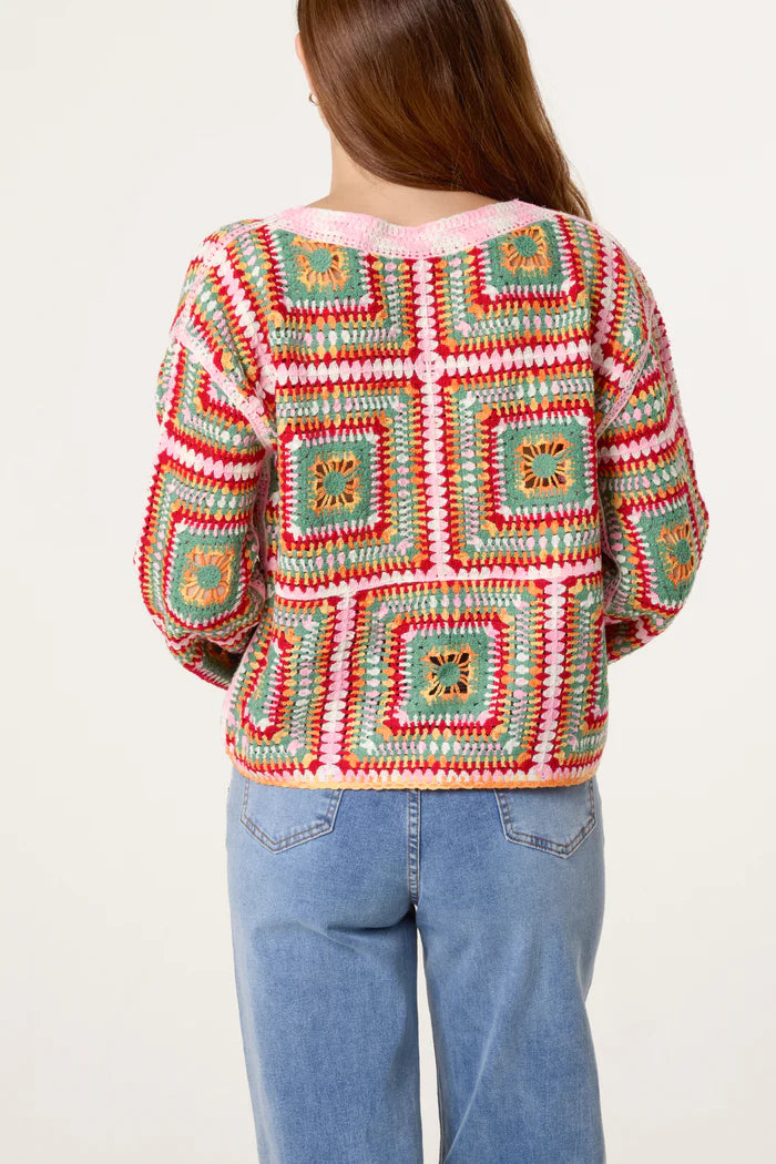 A person with long brown hair is seen from behind, wearing a rainbow crochet jumper with granny square patterns and light blue jeans.