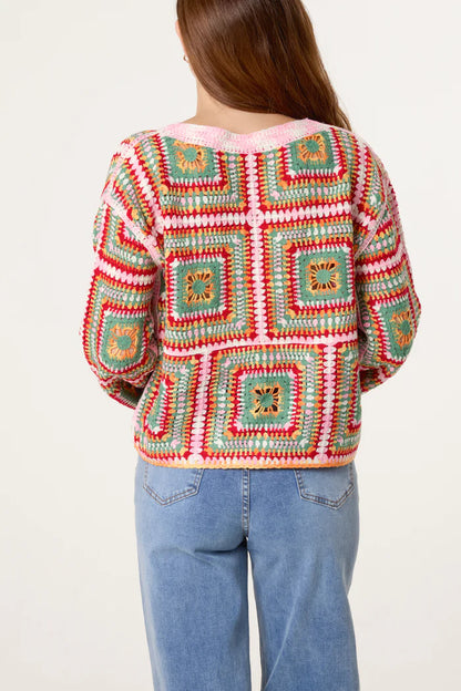 A person with long brown hair is seen from behind, wearing a rainbow crochet jumper with granny square patterns and light blue jeans.