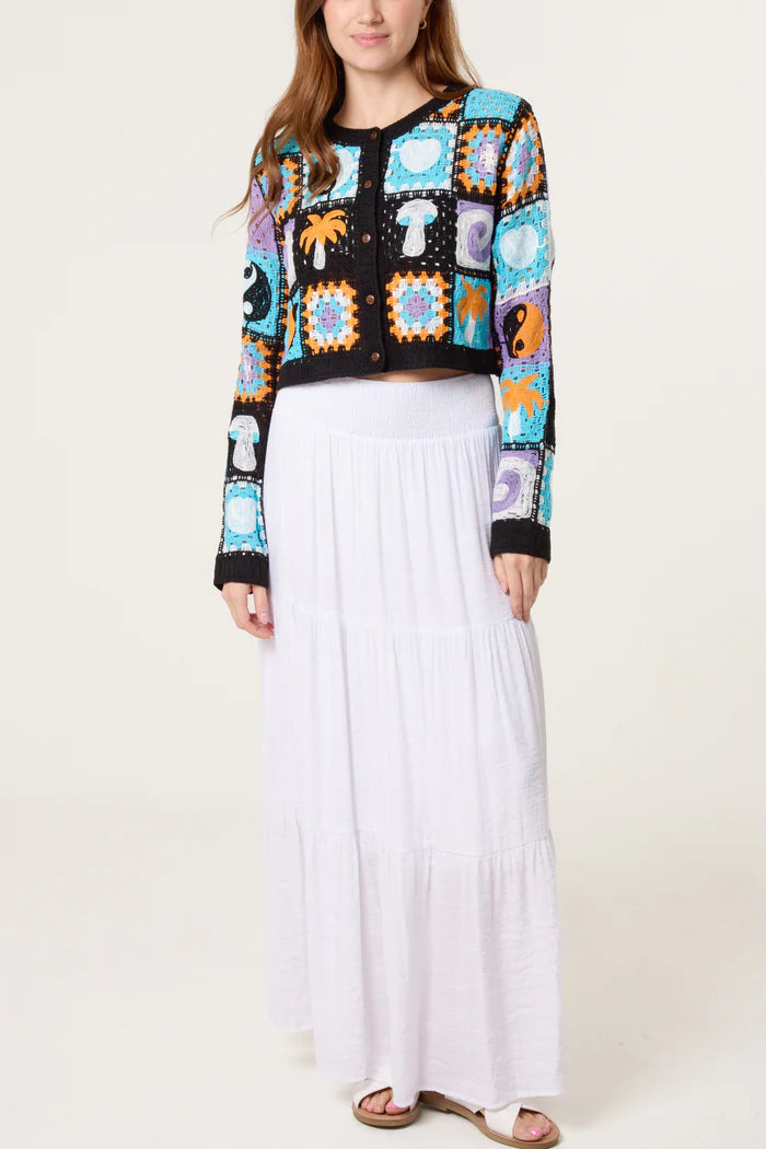 A woman in a white maxi skirt and a retro patchwork crochet cardigan featuring colourful square patterns and assorted motifs stands against a plain background.