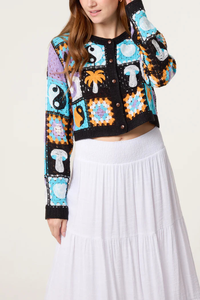 A woman dressed in a retro patchwork crochet cardigan featuring handcrafted crochet mushrooms, yin-yang symbols, and sun motifs, paired with a white tiered skirt.
