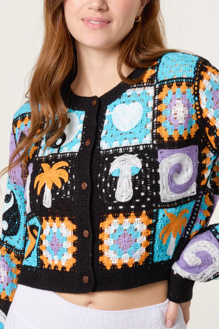 A woman wearing a retro patchwork crochet cardigan decorated with colourful designs such as a heart, mushroom, palm tree, swirl, and yin-yang symbols.