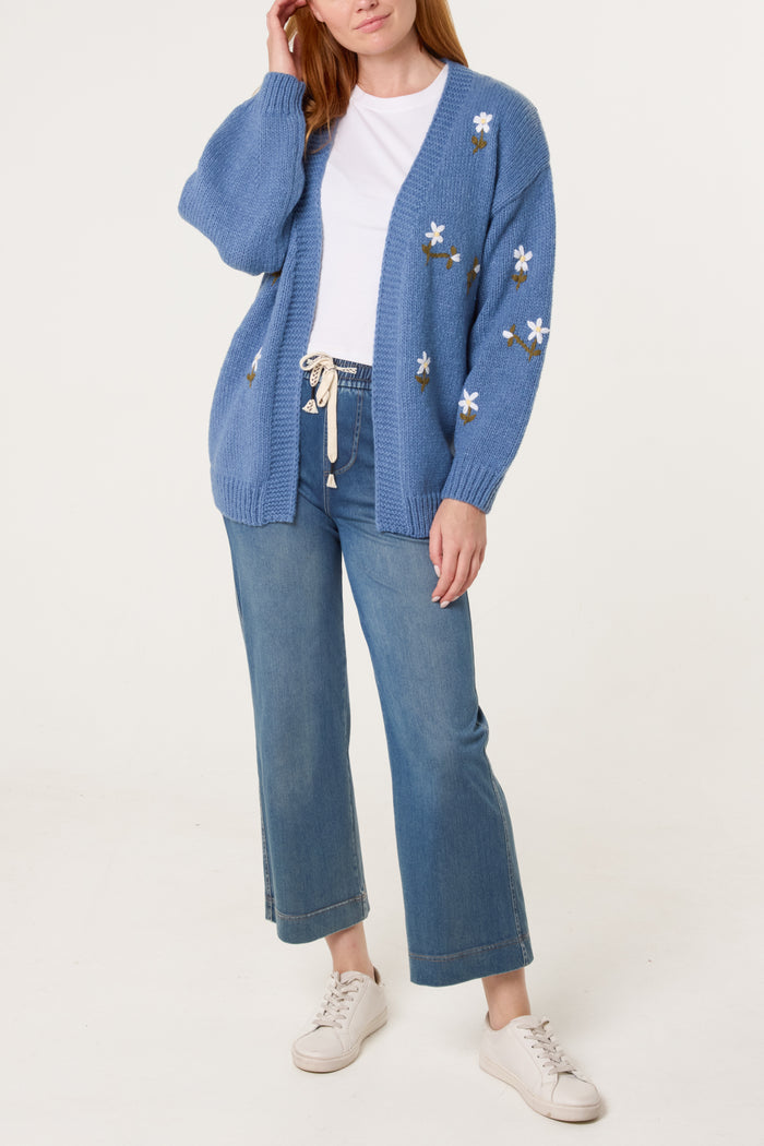 A person wearing a blue knitted cardigan with daisy embroidery, a white t-shirt, blue jeans, and white trainers stands against a plain white background.