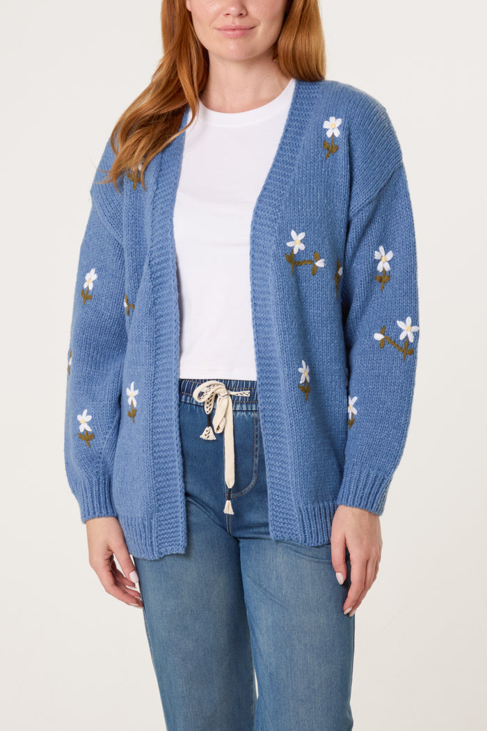 A person is wearing a blue knitted cardigan with daisy embroidery over a white T-shirt and blue jeans.