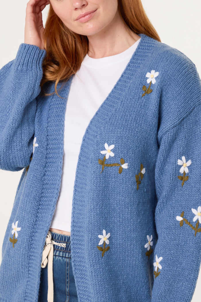 A person wearing a blue knitted cardigan with daisy embroidery over a white t-shirt and blue jeans.