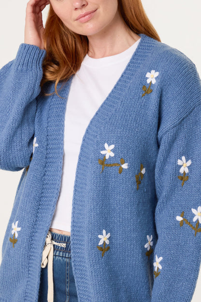 A person wearing a blue knitted cardigan with daisy embroidery over a white t-shirt and blue jeans.