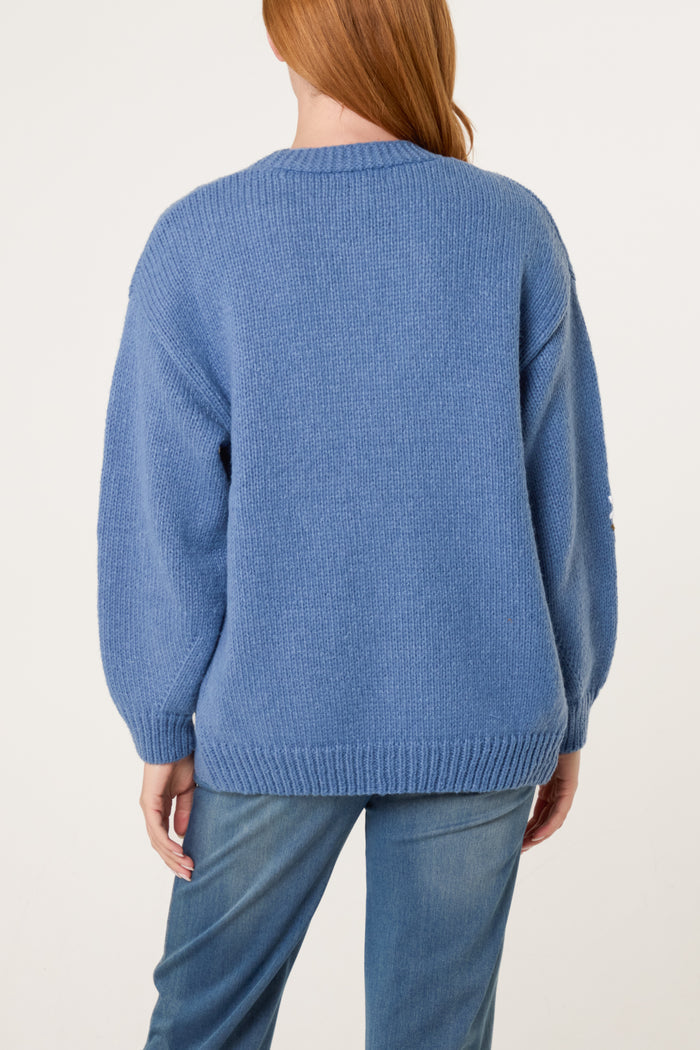 Person with long red hair wearing a blue knitted cardigan featuring daisy embroidery and light blue jeans, seen from the back against a plain background.