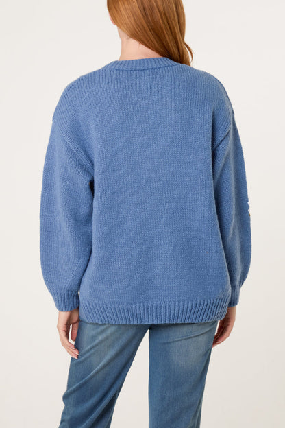 Person with long red hair wearing a blue knitted cardigan featuring daisy embroidery and light blue jeans, seen from the back against a plain background.