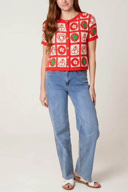 A person wearing a red and white crochet cardigan adorned with heart, flower, and fruit motifs, light blue jeans, and white sandals stands against a plain background.