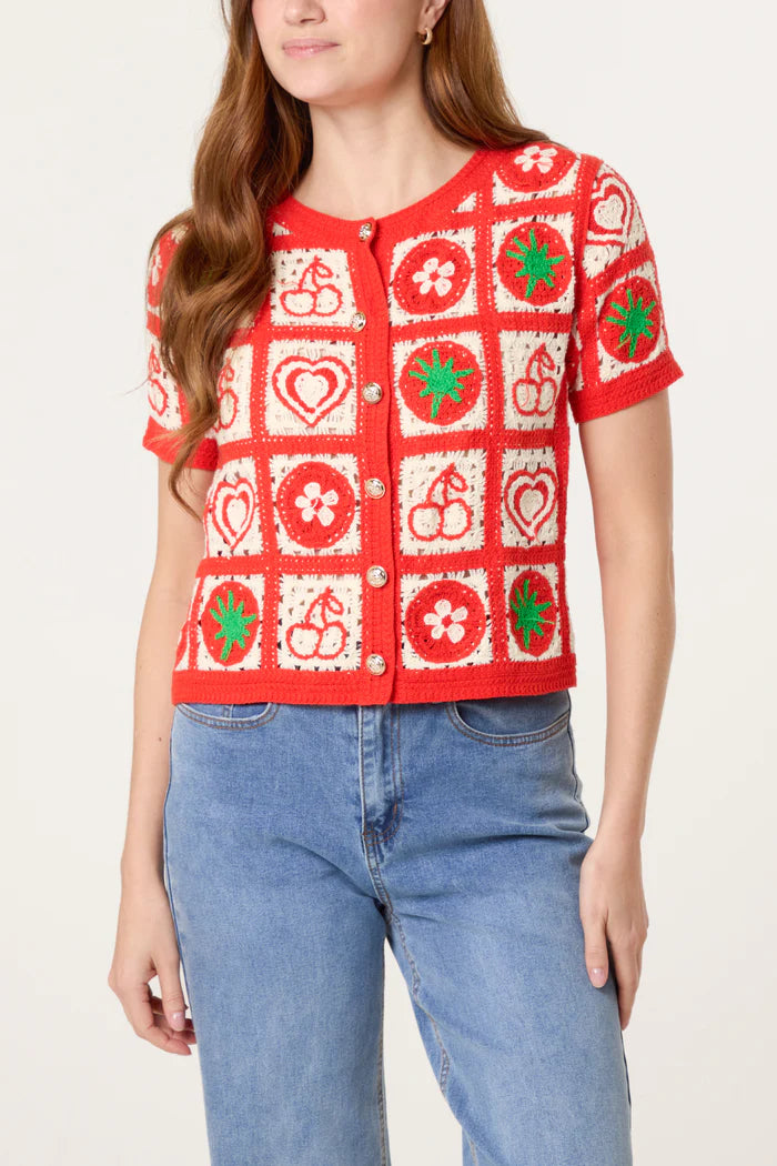 A person wearing a short-sleeved, red and white crochet cardigan adorned with playful fruit motifs and floral patterns, paired with blue jeans.
