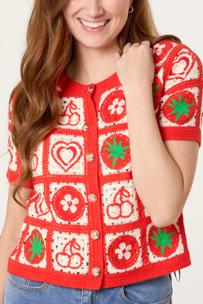 A woman dressed in a red crochet cardigan featuring cream and green fruit motifs and heart patterns in an appealing patchwork design, teamed with blue jeans.