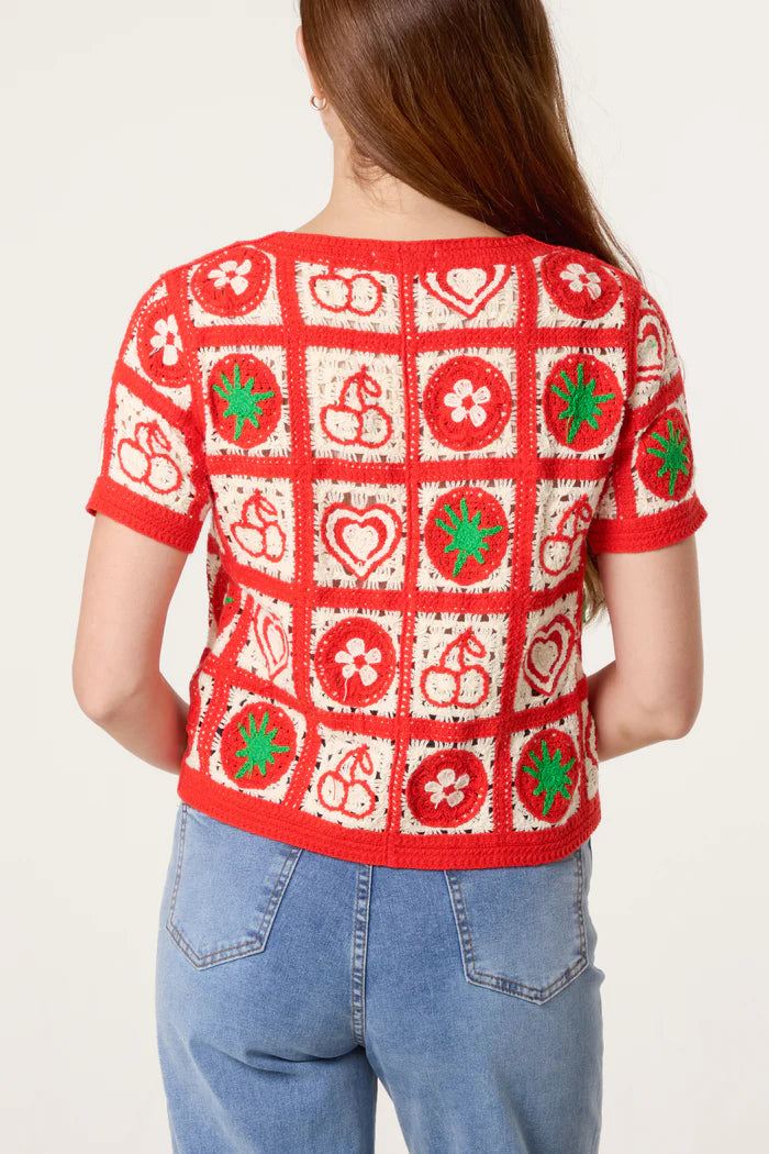 A person with long brown hair is seen from behind, wearing a red and white crochet cardigan adorned with fruit motifs and floral patterns, paired with blue jeans.