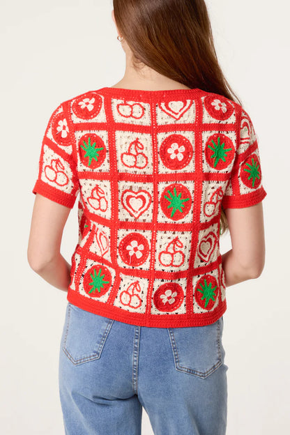 A person with long brown hair is seen from behind, wearing a red and white crochet cardigan adorned with fruit motifs and floral patterns, paired with blue jeans.